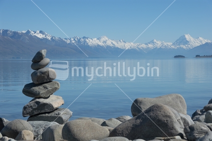 Stone stacks on shore of Lake Pukaki with Southern Alps and Mount Cook in distance across lake.