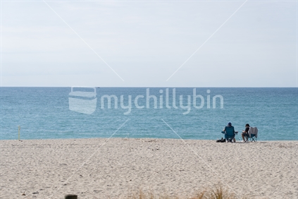 Tauranga New Zealand - August 25 2023- Two men sitting indistance on beach with water and sky.