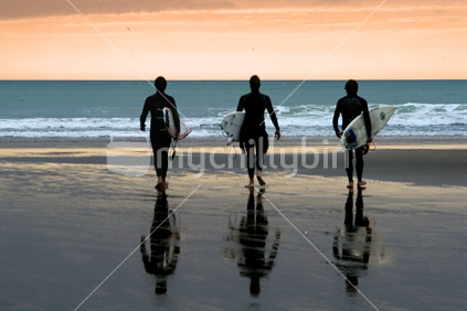 Three surfers walking towards the waves at Muriwai Beach