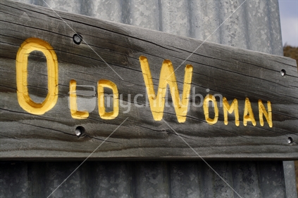A wooden sign with Old Woman carved in Gold lettering, attached to a corrugated iron fence.