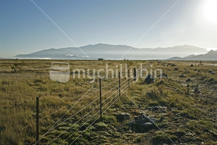 The plains of McKenzie basin, with misty hills and early morning sunlight accentuating wires on a boundary fence.