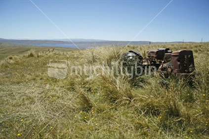 The body of a rusted old tractor lies amongst tussock in the high country, with Lake Onslow in the distance.  Central Otago.