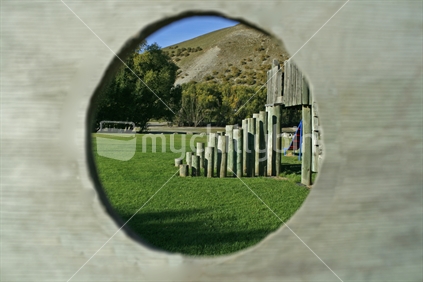 A children's playground seen through a hole in a wooden climbing frame .