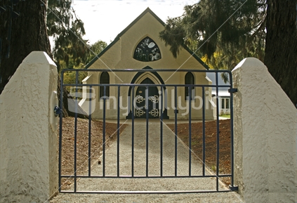 A white stucco church with unusual shaped window, behind a closed gate. Central Otago.