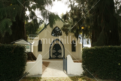 A white stucco church with unusual shaped window, set between huge redwood trees, Central Otago.