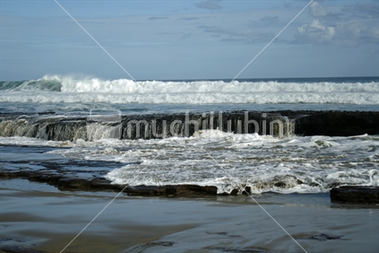 Ocean waves rolling over flat rocks onto a beach in Northland.