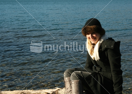 A young lady smiling, sitting on a log beside a lake, Central Otago.