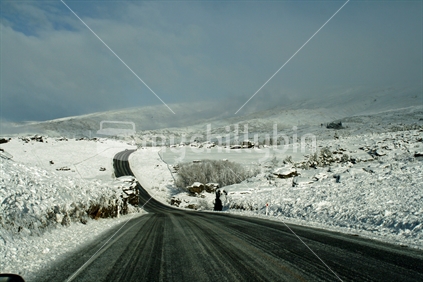 A black tarsealed road leading through farmland heavily blanketed with snow, in Central Otago.