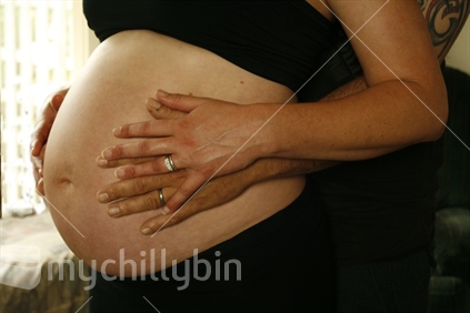 A mature woman in her third trimester gently holding her unborn child with her husband.