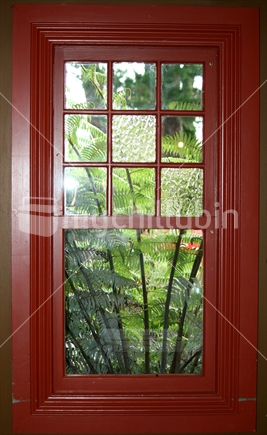 View through an old red wooden frame, of punga leaves in a garden
