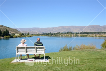 An elderly couple seated on a wooden bench, overlooking the Kawarau arm of Lake Dunstan, Central Otago.