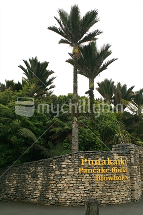 Public signage at the entrance of the walkway to Punakaiki rocks, with mature nikau palms and native bush, on a stormy overcast day
