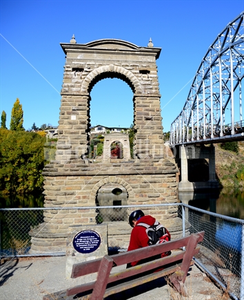 A cyclist seated resting overlooking the Historic stone supports of Alexandra bridge, alongside new steel one, both spanning Clutha River, Alexandra.