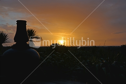 A chiminea fireplace surrounded by flax, silhouetted by a golden sunset. 