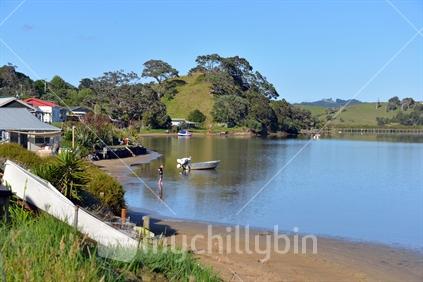 A child plays safely in the water of  a tidal estuary, surrounded by baches and boats, a typical Kiwi holiday.