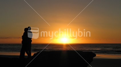 A couple hugging and watching the sun setting at a New Zealand beach.