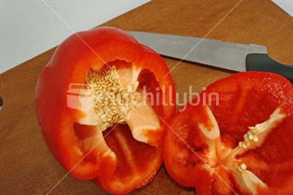 A fresh red bell pepper or capsicum and sharp knife on a wooden chopping board, cut on an angle exposing seeds and inside surface.