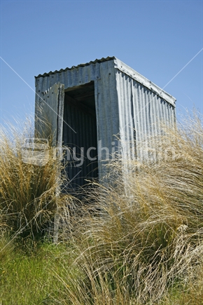 An old abandoned outhouse or toilet, surrounded by golden tussock grass, sits overlooking the lake.