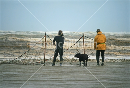 Two men and their dog, dragging a net on a wintery day, Oreti Beach, Southland.