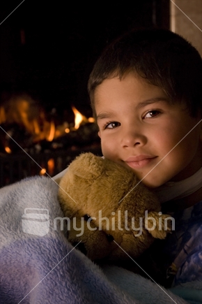 A boy cuddles a teddy in front of a fire