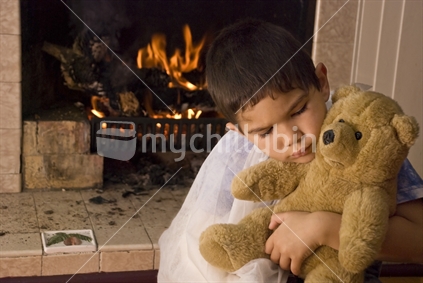 A boy with a broken arm cuddles a teddy in front of a fire