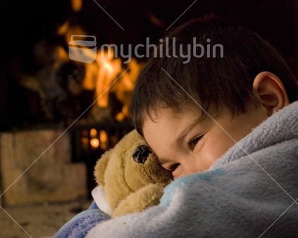 A boy cuddles a teddy in front of a warm fire