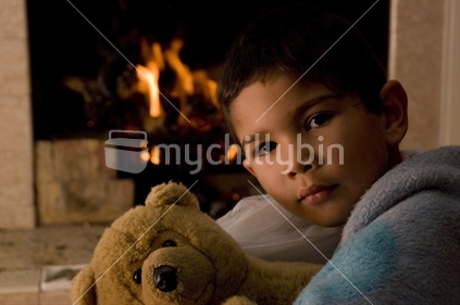 A curious looking boy sits in front of a fire, cuddling a teddy