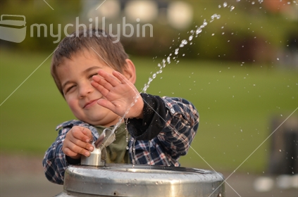 A boy sprays himself with water at a drinking fountain (focus hand)