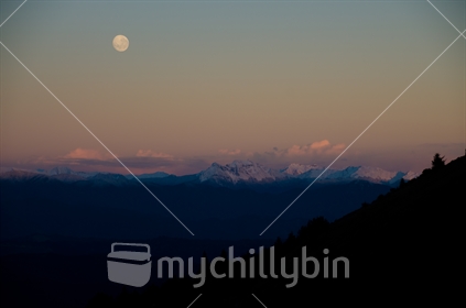 Full moon setting behind snow capped mountains