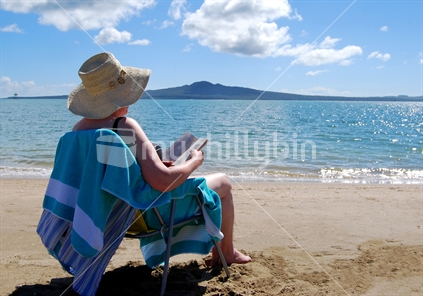 Elderly Lady sitting on Mission Bay beach, with view to Rangitoto Island, Auckland, New Zealand