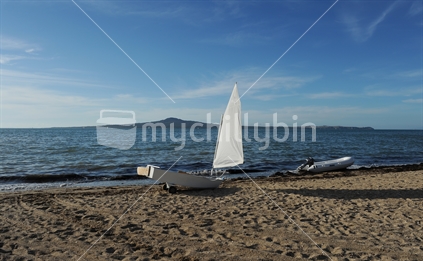 Optimist yacht with white sail on beach with Rangitoto island behind, New Zealand