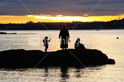 Silhouette of mother and daughters on rocks at Kohimarama beach at sunset, New Zealand