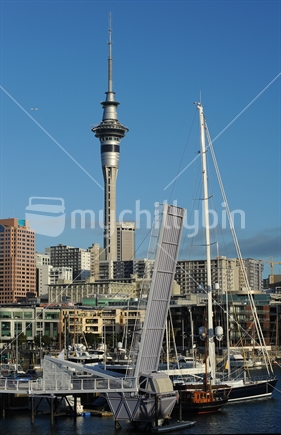 Yachts and Te Wero drawbridge at the Viaduct Basin, with the CBD, Skytower and jet in the background against a blue sky ; Auckland, New Zealand.  