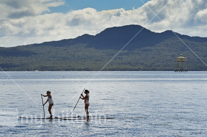 Two women on Paddle boards, with Rangitoto beyond.