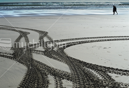 Tyre track patterns in west coast sand, with two people  silhouetted walking in the background. 