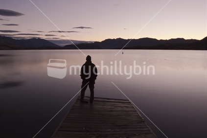 A man standing on an old jetty looking out over Lake Wanaka in the early morning light, New Zealand