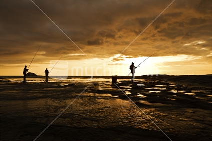 Three men fishing from the rocks at Muriwai, silhouetted against the setting sun. New Zealand