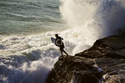 A surfer leaps from the rocks into rough water at Muriwai, New Zealand.