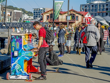 Wellington Waterfront near Frank Kitts Park on a fine Sunday afternoon with folks strolling and enjoying themselves. One man in a red shirt plays the piano. The focus is on the piano player