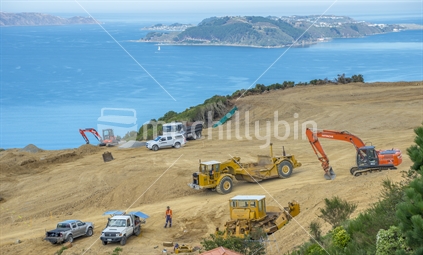 Earthworks for a small luxury housing subdivision being carried out on the hills  in Newlands, Wellington. Varous machines can be seen from excavators, trucks, bulldozers and a  scraper. In the distance the waters of Wellington Harbour