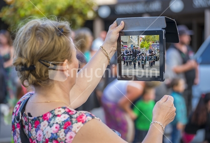 A middle aged woman films a piper band in a christma street parade, on a tablet device
