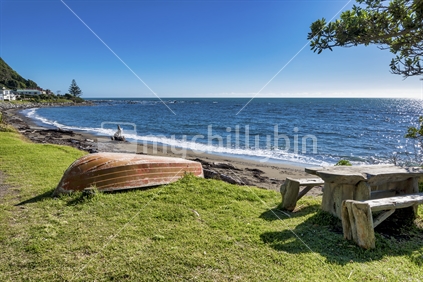 Pukerua bay on a fine sunny autumn day showing the grass verge, a row boat, a rustic picnic table and an inviting sea with small surf