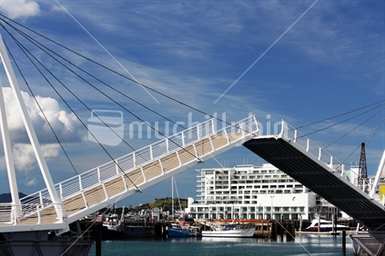 The new white bridge at Wynyard Quarter, Viaduct Harbour, in Auckland, lifting up framing a view of commercial fishing boats and the Hilton Hotel behind. 