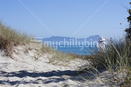 A woman in white clothes stands on Waipu Cove Beach sand dune, and looks out to Whangarei Heads, New Zealand.
