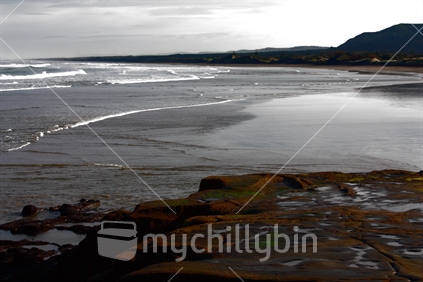 Foreground rock pools with a bleak winter view of deserted Muriwai Beach and silhouetted hills.