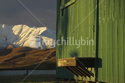 Wedderburn rail siding, Otago. Side of the corrugated iron wall with seat and shadows and snow covered mountain range in the background