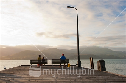 Mother and daughter share bench, and the pier with fisherman, in the early evening.
