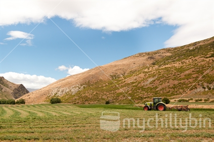Making hay while the sun shines; tractor mowing hay with hills and sunny blue sky in background.