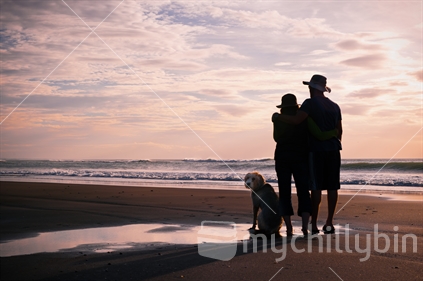 Young couple watching pink sunset from beach with their dog