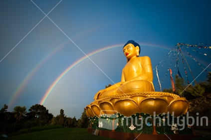 A golden statue of Buddha sits beneath a double rainbow, with New Zealand cabbage trees to the side. 